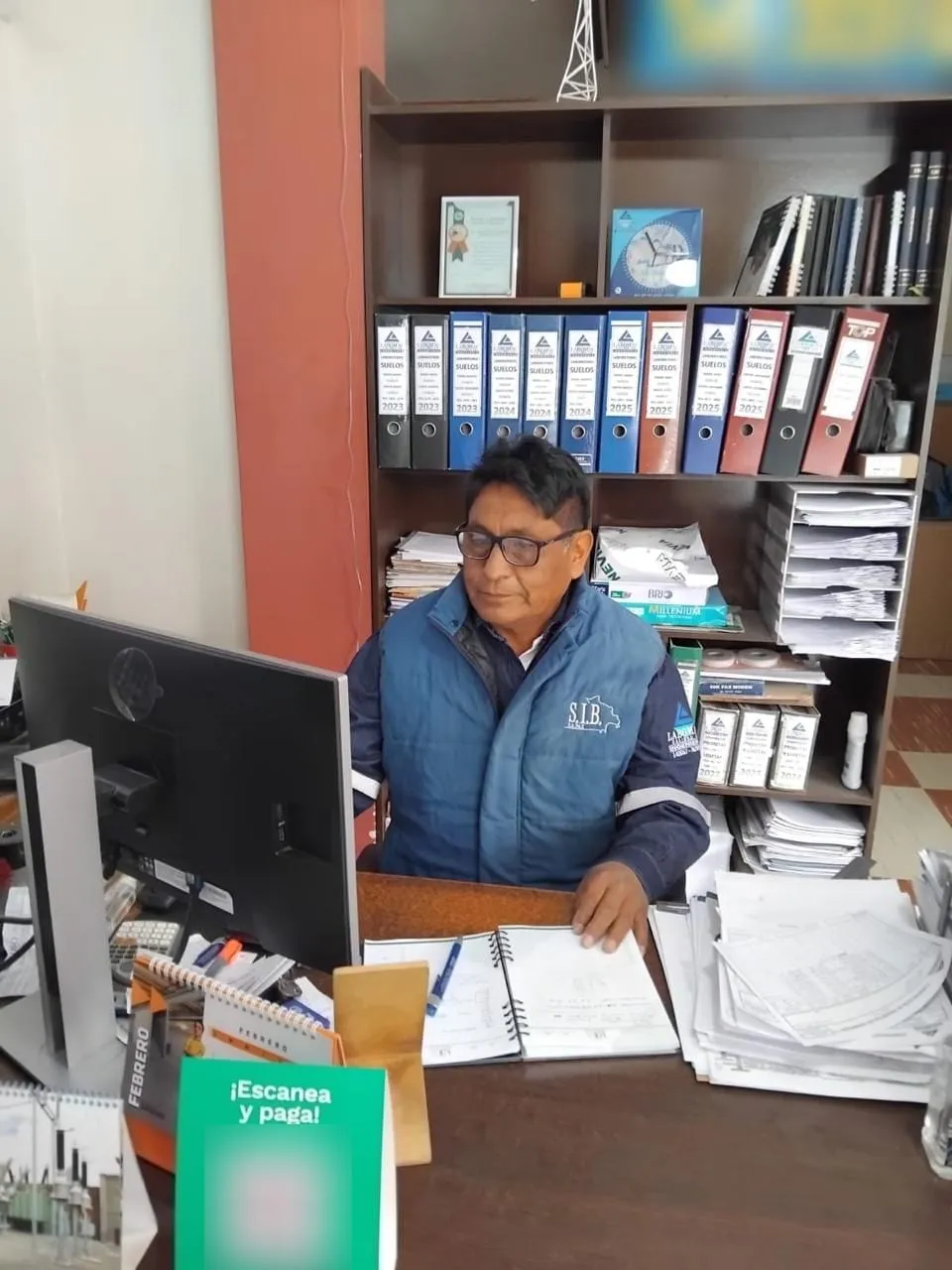 El Ing. Walter Castro, con chaleco azul, revisa documentos en su escritorio frente a estantes con archivadores.